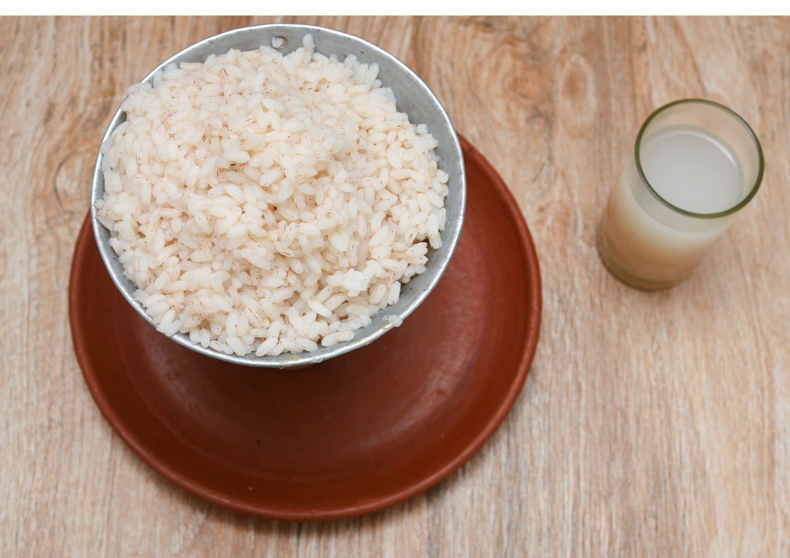 A bowl of cooked rice on a clay plate beside a glass of fermented rice water.